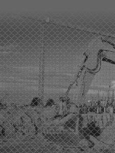 A monochrome image of a construction site with an excavator. The scene is viewed through a chain-link fence, partially obstructing the view. Several workers are present near the excavator, which appears to be digging into the ground.